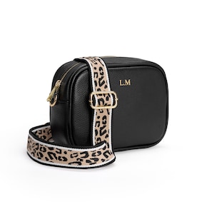 May include: A black pebbled leather camera bag featuring gold-tone hardware, a top zipper closure, and the gold-embossed initials L.M. on the front. The bag includes a wide, adjustable crossbody strap with a beige and black leopard print pattern.