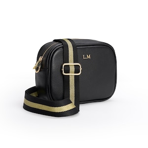May include: A black pebbled leather camera bag featuring gold-tone hardware, a top zipper closure, and the initials L.M embossed in gold. The bag includes an adjustable black and gold striped webbing shoulder strap with a gold metal slider buckle.