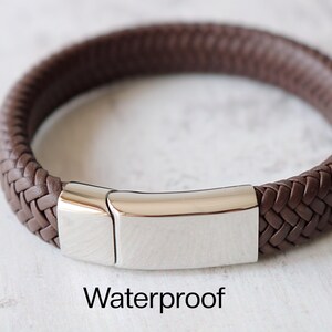 May include: A brown leather bracelet with a silver clasp. The bracelet is braided and has a smooth, polished finish. The clasp is a simple, rectangular design. The text "Waterproof" is printed below the bracelet.