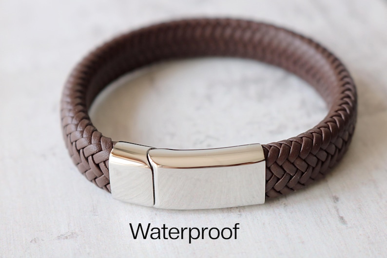 May include: A brown leather bracelet with a silver clasp. The bracelet is braided and has a smooth, polished finish. The clasp is a simple, rectangular design. The text "Waterproof" is printed below the bracelet.