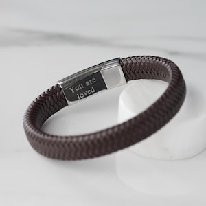 May include: A brown leather bracelet with a silver clasp. The clasp is engraved with the words "You are loved".