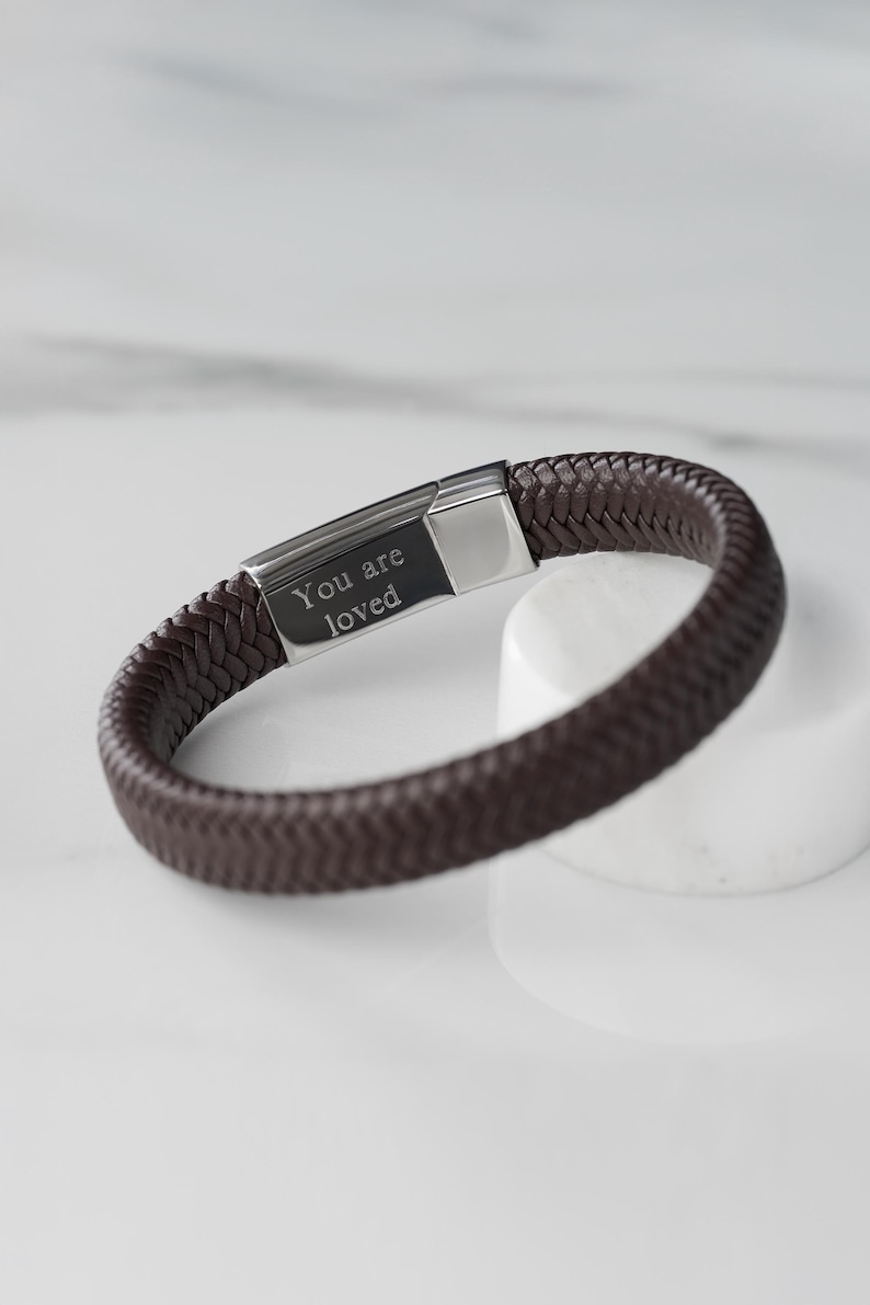 May include: A brown leather bracelet with a silver clasp. The clasp is engraved with the words "You are loved".