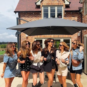 Peut inclure: Six personnes portant diverses tenues, notamment des robes en jean, des shorts noirs et des combishorts beiges, chacune accessoiris&eacute;e avec un sac banane en bandouli&egrave;re. Elles portent des bottes noires et des lunettes de soleil sous un grand parasol gris.