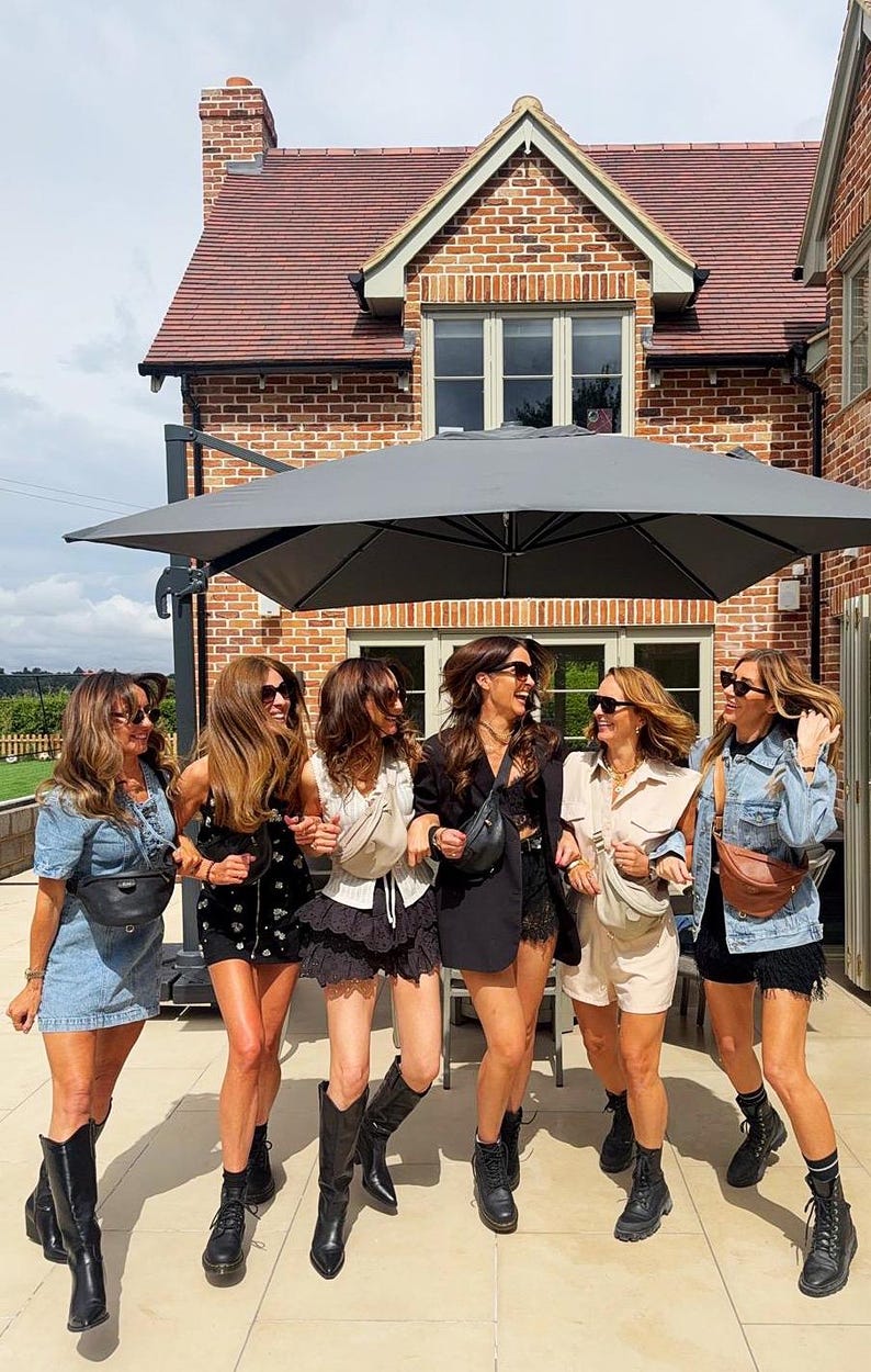 Peut inclure: Six personnes portant diverses tenues, notamment des robes en jean, des shorts noirs et des combishorts beiges, chacune accessoiris&eacute;e avec un sac banane en bandouli&egrave;re. Elles portent des bottes noires et des lunettes de soleil sous un grand parasol gris.