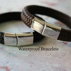 Op de afbeelding: Twee gevlochten lederen armbanden met zilveren magneetsluitingen. De armbanden zijn bruin en zwart. De tekst "Waterdichte armbanden" is zichtbaar onder de armbanden.