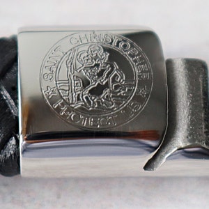 Op de afbeelding: Een zilveren armband met een zwarte leren band en een zilveren bedel met de woorden "Sint Christoffel Bescherm Ons" gegraveerd.