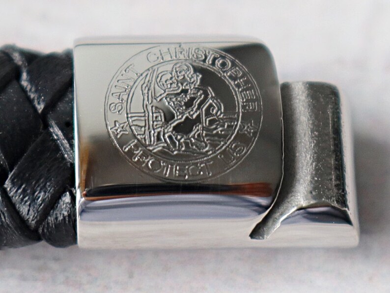 Op de afbeelding: Een zilveren armband met een zwarte leren band en een zilveren bedel met de woorden "Sint Christoffel Bescherm Ons" gegraveerd.