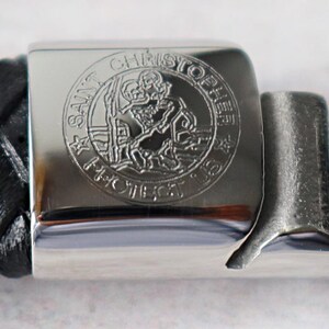 May include: A silver metal bracelet clasp with a black leather strap. The clasp features an engraved image of Saint Christopher carrying a child, with the words "Saint Christopher Protect Us" around the image.