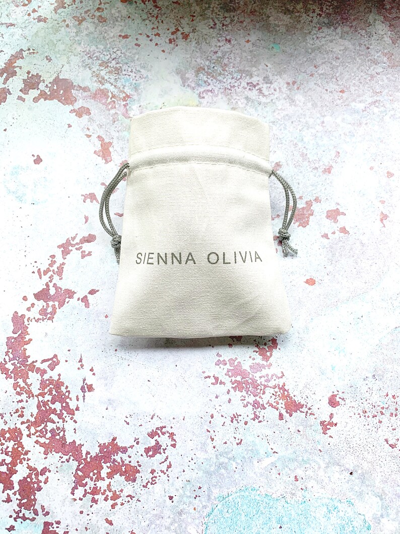 Op de afbeelding: Een witte trekkoordtas met de tekst "SIENNA OLIVIA" gedrukt op de voorkant.