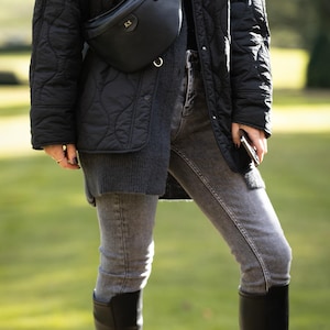 Peut inclure: Une veste noire matelass&eacute;e port&eacute;e sur un pull gris fonc&eacute; et un jean gris, associ&eacute;e &agrave; des bottes noires arrivant au genou et une sacoche banane en cuir noir avec les initiales KS dor&eacute;es en relief et un anneau en m&eacute;tal dor&eacute;.