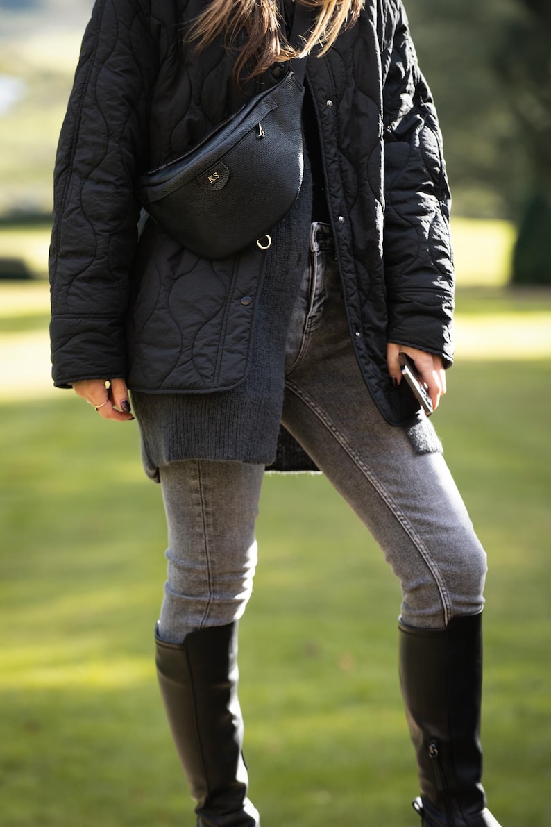 Peut inclure: Une veste noire matelass&eacute;e port&eacute;e sur un pull gris fonc&eacute; et un jean gris, associ&eacute;e &agrave; des bottes noires arrivant au genou et une sacoche banane en cuir noir avec les initiales KS dor&eacute;es en relief et un anneau en m&eacute;tal dor&eacute;.