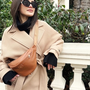 Peut inclure: Un sac bandouli&egrave;re en cuir marron clair &agrave; texture grain&eacute;e, dot&eacute; d'une fermeture &eacute;clair dor&eacute;e et d'une sangle r&eacute;glable assortie. Le sac est port&eacute; sur un manteau en laine beige avec un pull &agrave; col roul&eacute; noir et des lunettes de soleil ovales.