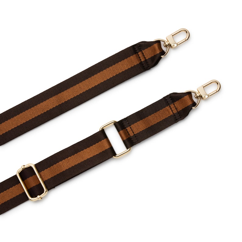 May include: Two adjustable bag straps featuring a dark brown and copper-colored striped pattern. Each strap is constructed from woven fabric and includes gold-toned metal hardware, specifically lobster claw clasps and rectangular sliders.