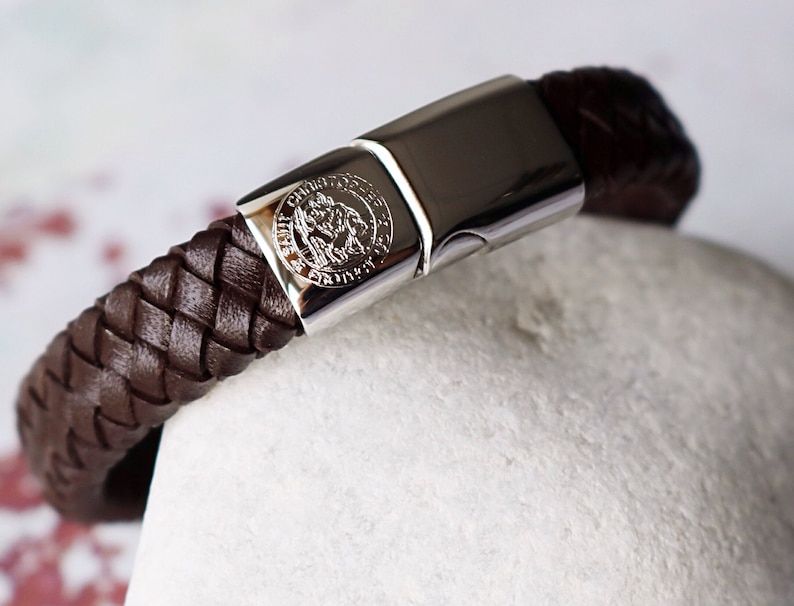 May include: A brown leather bracelet with a braided design and a silver clasp. The clasp features a circular design with an embossed image.