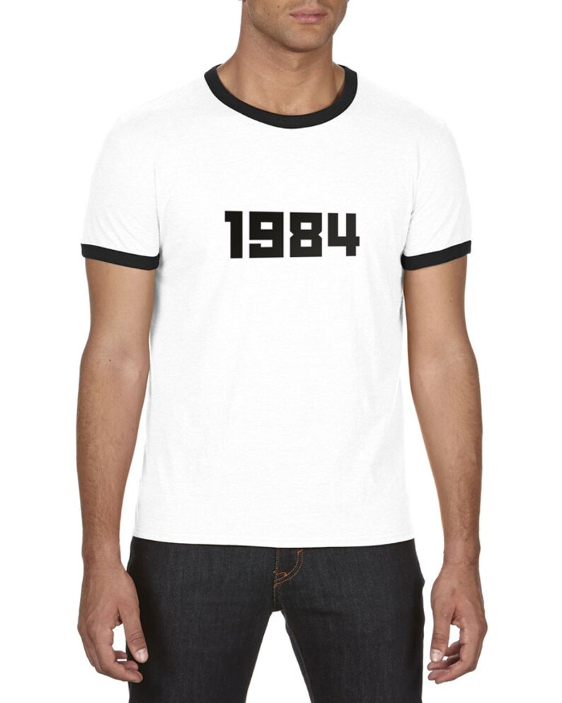 May include: White t-shirt with a black ringer and the number 1984 printed on the front.