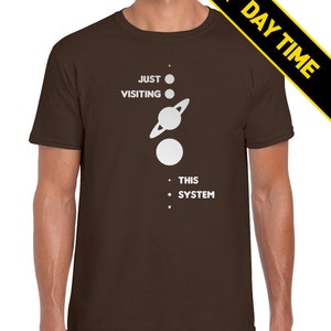 May include: A brown t-shirt with a white graphic of a solar system with the text 'JUST VISITING THIS SYSTEM'.