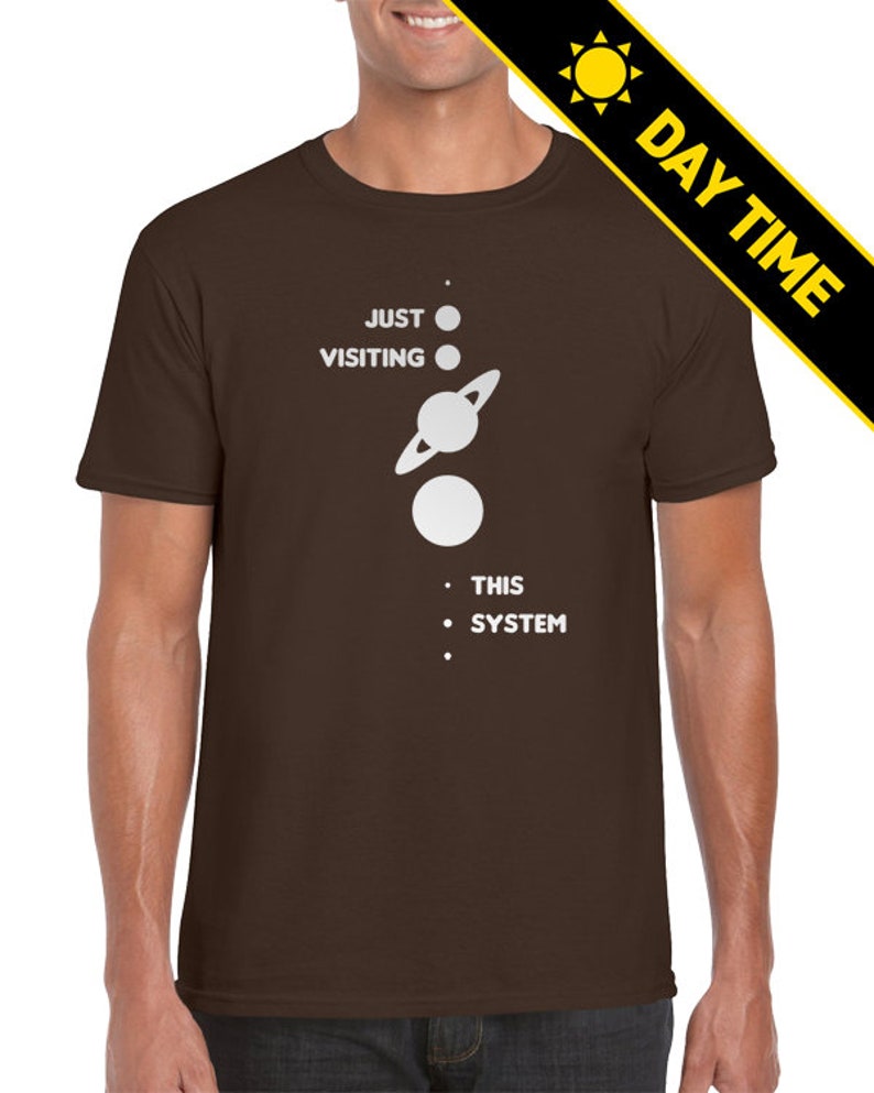 May include: A brown t-shirt with a white graphic of a solar system with the text 'JUST VISITING THIS SYSTEM'.