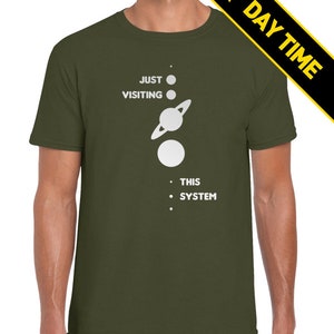 May include: A green t-shirt with white text that reads 'Just Visiting This System' and a graphic of a solar system with planets and a sun.