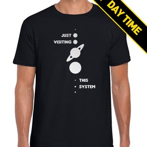 May include: A black t-shirt with a white graphic of a solar system with the text 'JUST VISITING THIS SYSTEM'.