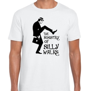 Puede incluir: Camiseta blanca con una silueta negra de un hombre con sombrero caminando con un maletín. El texto "The Ministry of Silly Walks" está impreso debajo de la silueta.