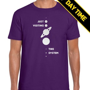 May include: A purple t-shirt with a white graphic of a solar system with the text 'JUST VISITING THIS SYSTEM'.