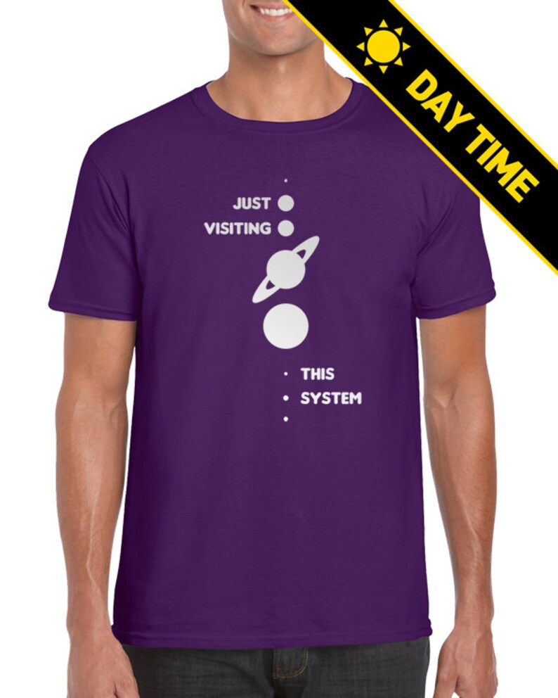 May include: A purple t-shirt with a white graphic of a solar system with the text 'JUST VISITING THIS SYSTEM'.