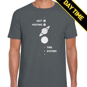 May include: A dark gray t-shirt with a white graphic of a solar system with the text 'JUST VISITING THIS SYSTEM'.