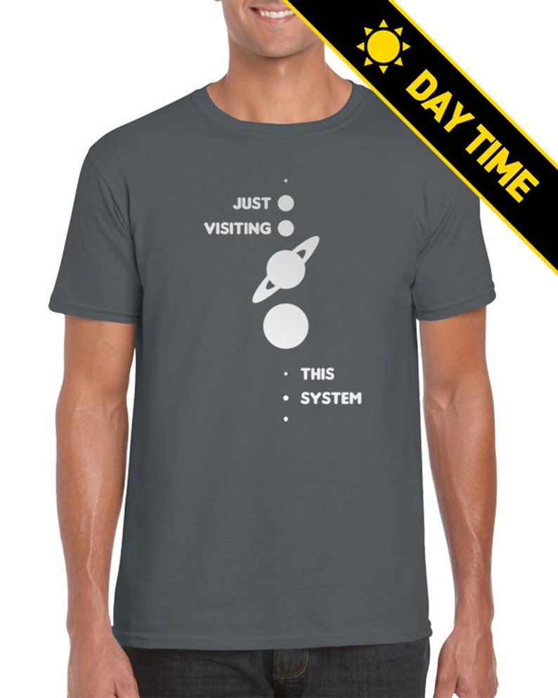 May include: A dark gray t-shirt with a white graphic of a solar system with the text 'JUST VISITING THIS SYSTEM'.