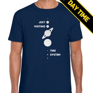 May include: A navy blue t-shirt with a white graphic of a solar system with the text 'JUST VISITING THIS SYSTEM'.
