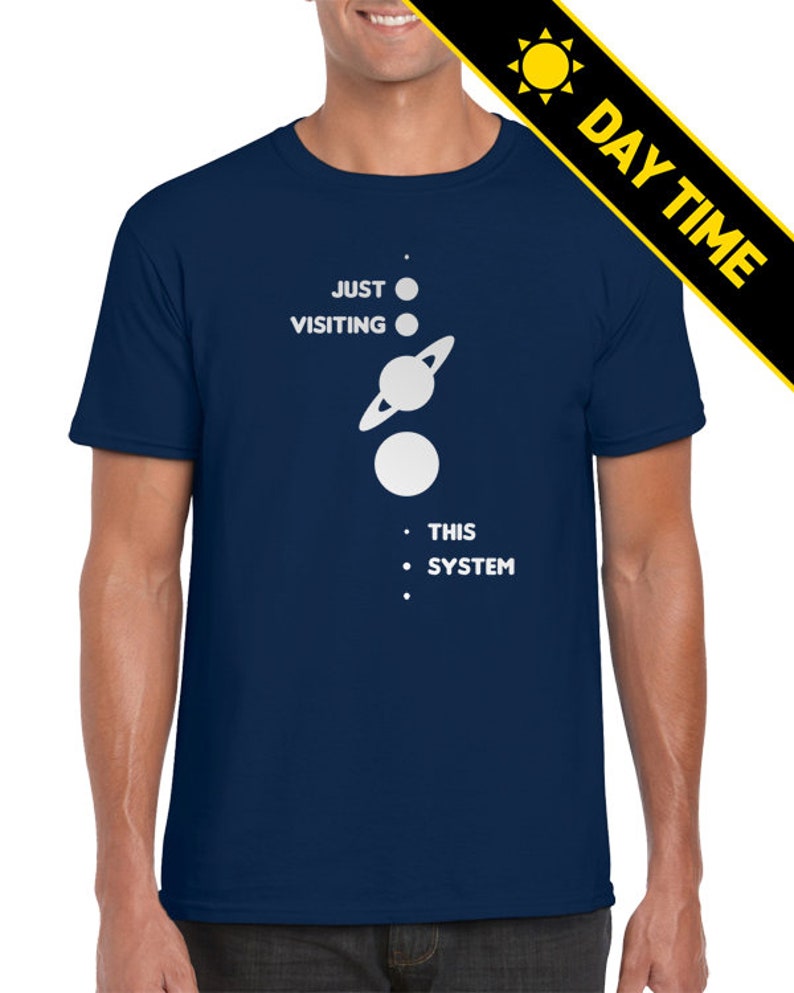 May include: A navy blue t-shirt with a white graphic of a solar system with the text 'JUST VISITING THIS SYSTEM'.