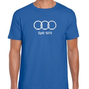 Può includere: Una t-shirt blu reale con una grafica bianca degli anelli olimpici con gocce d'acqua sotto. Il testo "Split 1979" è sotto gli anelli.