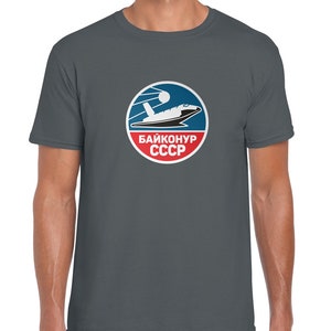 Pode incluir: Uma camiseta cinza escura com um logotipo circular que apresenta uma nave espacial branca, um planeta azul e uma estrela vermelha. O logotipo também inclui o texto "БАЙКОНУР СССР" em branco.