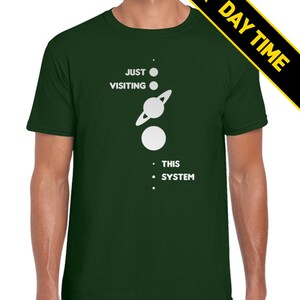 May include: A dark green t-shirt with a white graphic of a solar system with the text 'JUST VISITING THIS SYSTEM'.