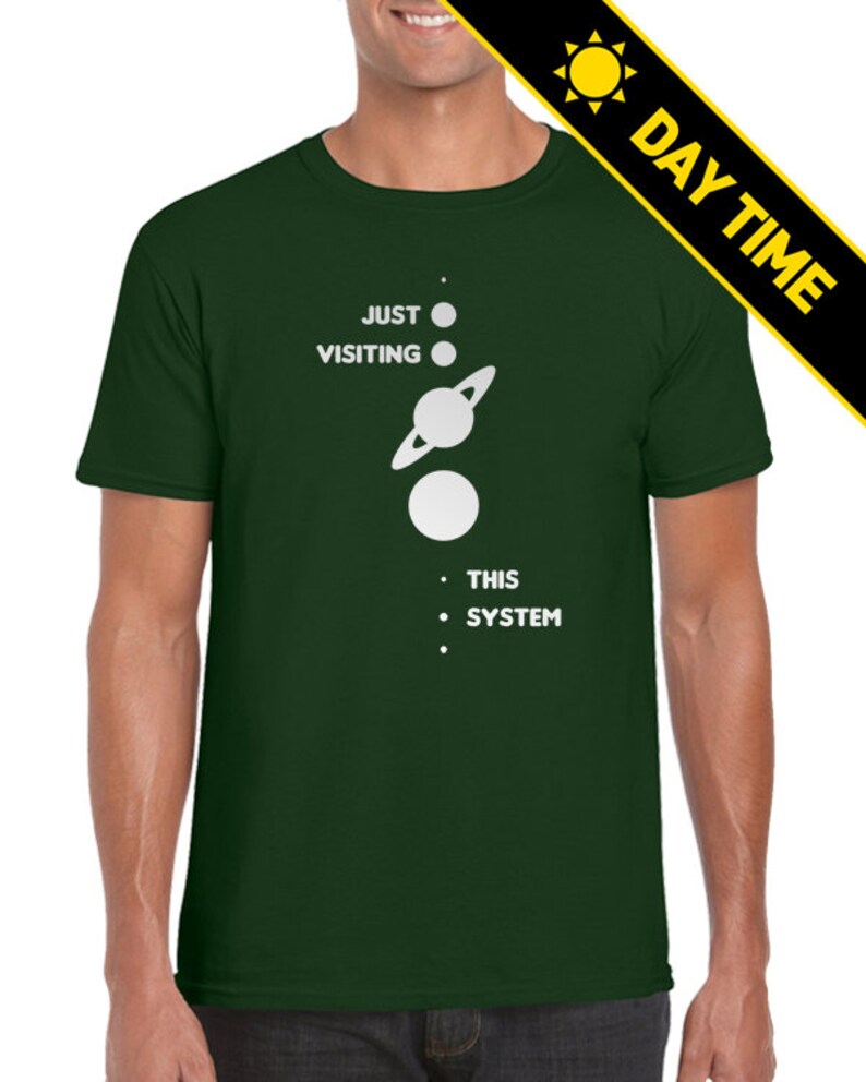 May include: A dark green t-shirt with a white graphic of a solar system with the text 'JUST VISITING THIS SYSTEM'.