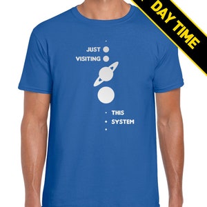 May include: A royal blue t-shirt with a white graphic of a planet with rings and three white dots. The text on the t-shirt reads 'Just Visiting This System'.