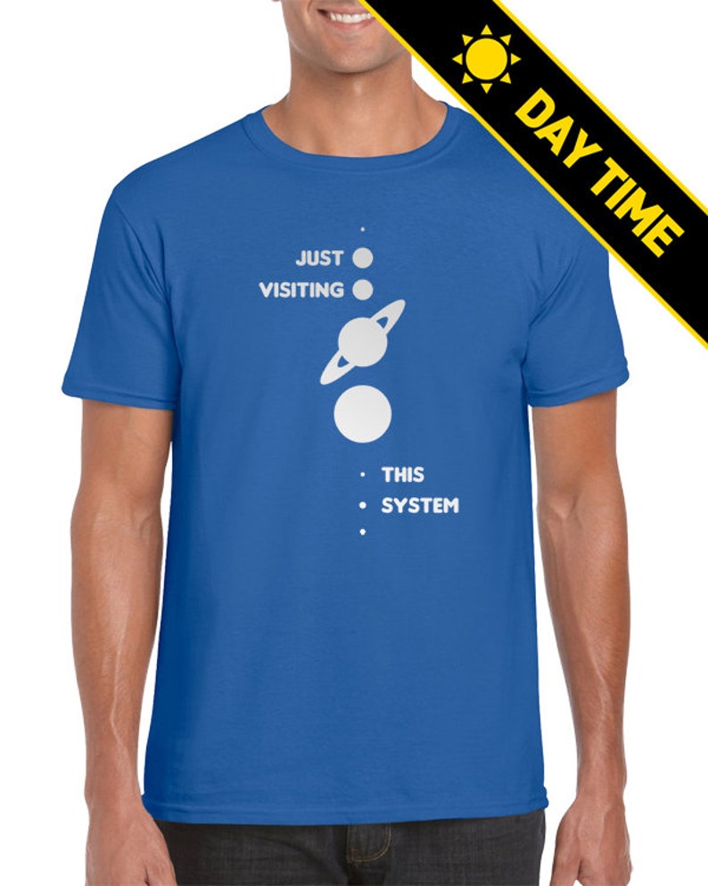 May include: A royal blue t-shirt with a white graphic of a planet with rings and three white dots. The text on the t-shirt reads 'Just Visiting This System'.