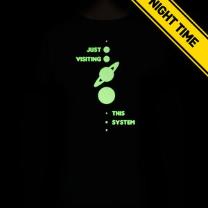 May include: A black t-shirt with a glow-in-the-dark design of a solar system with the text 'Just Visiting This System'.