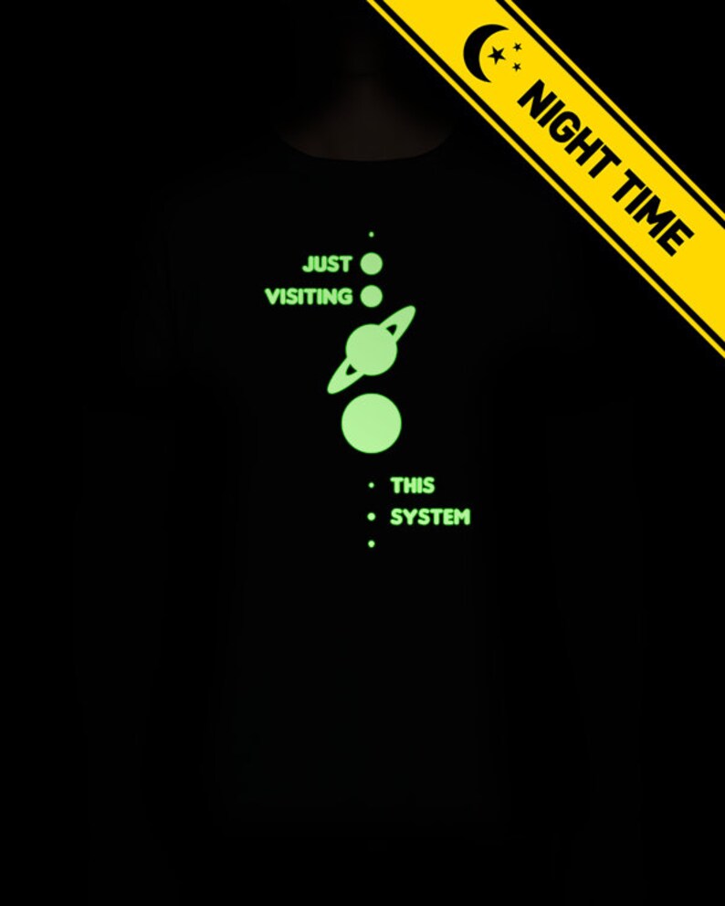 May include: A black t-shirt with a glow-in-the-dark design of a solar system with the text 'Just Visiting This System'.
