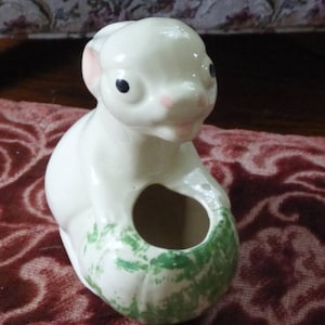 Puede incluir: Figura de ardilla de cerámica color crema, con detalles rosas en las orejas y la nariz, y ojos negros. La ardilla sostiene una maceta pintada de verde y blanco. La figura es probablemente vintage y decorativa.