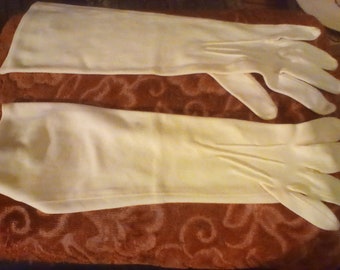 May include: A pair of long, cream-coloured gloves. The gloves are made of a smooth material. The gloves extend past the wrist, covering a portion of the forearm. The gloves are laid out on a patterned, reddish-brown surface.