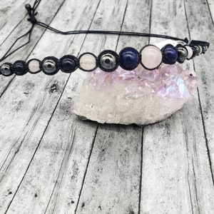 May include: A black cord bracelet with blue, pink, and grey beads. The bracelet is on a white, textured surface.