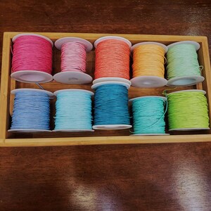Peut inclure: Dix bobines de fil &agrave; broder color&eacute; dans une bo&icirc;te en bois. Les couleurs sont rose, rose clair, orange, jaune, vert clair, bleu, bleu clair et vert.