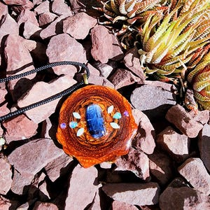 May include: A handcrafted pendant necklace featuring a central blue stone, surrounded by smaller white, blue, and purple stones, set in a brown, wood-like resin. The pendant hangs from a black cord. The background is composed of reddish-brown rocks and green and yellow succulents.