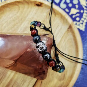 Peut inclure: Un bracelet en cordon noir avec des perles de pierres pr&eacute;cieuses color&eacute;es et un charme Bouddha argent&eacute;. Le bracelet est sur un cristal rose.