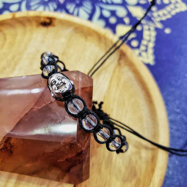 Peut inclure: Un bracelet noir &agrave; perles avec un charme en argent repr&eacute;sentant un visage de Bouddha souriant. Le bracelet est sur une surface rose et brune.