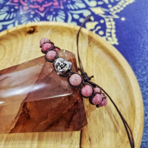 Peut inclure: Un bracelet en perles roses et brunes avec un charme argent&eacute; repr&eacute;sentant un visage souriant. Le bracelet est sur une surface en bois.