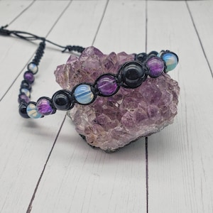 May include: A black cord bracelet with purple, blue, and black beads. The bracelet is resting on a purple amethyst crystal cluster.