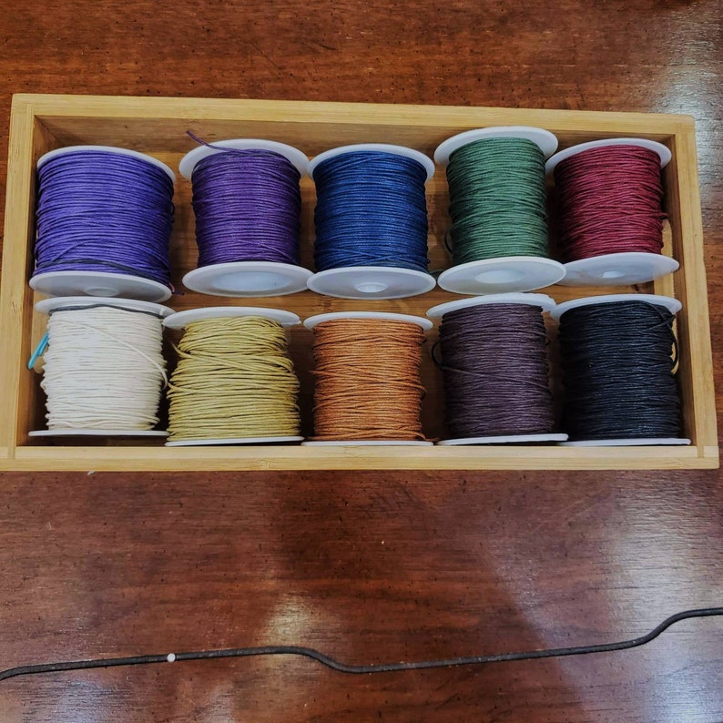 Peut inclure: Une bo&icirc;te en bois remplie de bobines de cordon cir&eacute; en coton de diff&eacute;rentes couleurs. Les cordons sont dans des tons de violet, bleu, vert, rouge, blanc, jaune, marron, noir et gris.