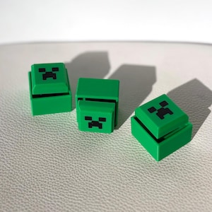 May include: Three green, cube-shaped objects with black facial features, resembling a popular video game character. The objects are arranged on a light-coloured surface, with shadows suggesting a light source from above.