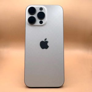 May include: A silver smartphone with a matte finish featuring a centered black Apple logo. The device has a rectangular camera module in the upper left corner containing three circular black camera lenses and a small circular flash component.